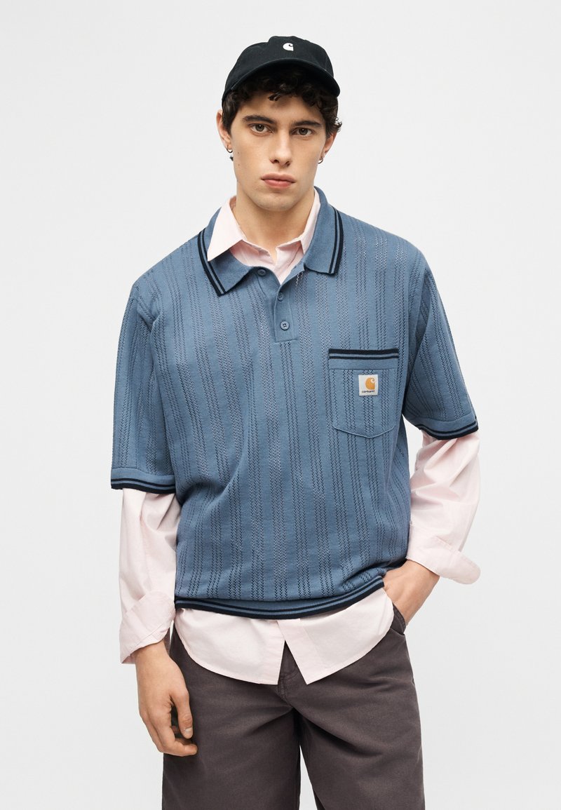 Jonge man die een blauw gebreid poloshirt draagt over een lichtroze overhemd, donkere broek en een zwarte pet, staand met één hand in de zak.