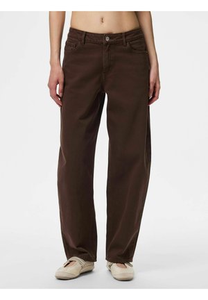 Person iført mørkebrune løstsiddende jeans og lysebeige slip-on sko, står mod en ensfarvet hvid baggrund.