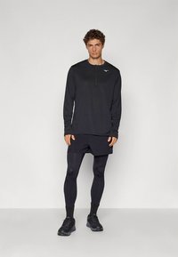 Zwart atletisch shirt met lange mouwen en halve rits, gecombineerd met donkere korte broeken en leggings. Zwarte sportschoenen en logodetail op het shirt.