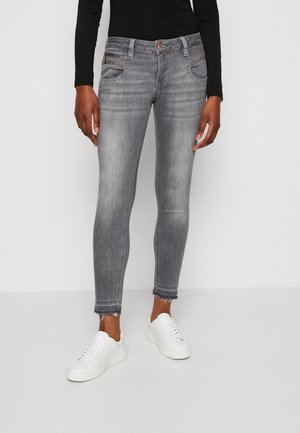 Person iført falmede grå skinny jeans med pyntede forlommer og frynsete kanter, kombineret med hvide sneakers og en sort langærmet top.
