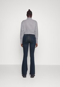 Mujer con cabello trenzado que lleva una chaqueta a cuadros en blanco y negro, jeans acampanados oscuros y zapatos negros, de pie contra un fondo liso.