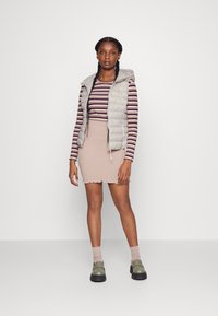 Hellgraue Pufferweste über einem gestreiften Langarmoberteil. Beige gerippter Minirock mit gewelltem Saum, kombiniert mit grünen Schuhen und hellen Socken.