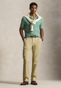 Mężczyzna stoi z rękami w kieszeniach, ubrany w jasnozieloną, krótkim rękawie koszulkę polo, kremowy sweter zarzucony na ramiona, beżowe spodnie i brązowe buty.