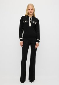 Maglione nero con collo a camicia, che presenta strisce verticali a contrasto in crema e bottoni. Abbinato a pantaloni neri a maniche svasate aderenti.
