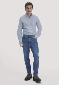 Man staat met één hand in zijn zak, draagt een lichtblauw knoopshirt, blauwe jeans en bruine veterboots op een effen achtergrond.