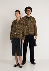 Dva modely v zladených textúrovaných hnedých sakoch na gombíky; jeden v čiernej koženej sukni a balerínach, druhý v čiernych nohaviciach a topánkach, ktorý drží tašku.