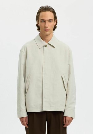 Jonge man met halflang haar draagt een lichtbeige jas met kraag over een donkere broek, staand tegen een effen lichte achtergrond.