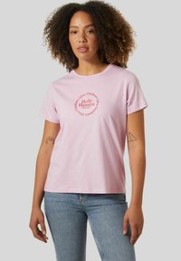 Camiseta de algodón rosa con un gráfico redondo que presenta "Helly Hansen" y texto relacionado con actividades al aire libre en rojo. Mangas cortas y corte relajado.
