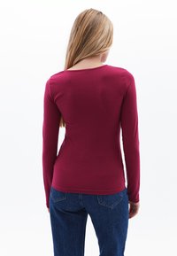T-särk, millel on pikad varrukad, burgundi värvi ning ümar kaelus, sile tekstuur ja liibuv lõige, kombineeritud tumedate siniste teksadega.
