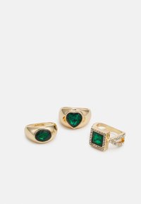 Três anéis de ouro com pedras preciosas verdes: dois em forma oval e um em forma de coração, além de um anel quadrado com pedras claras ao longo da banda.