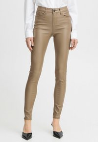Pantalons taille haute, ajustés, en matériau lisse de couleur beige avec un léger brillant. Présentent un design classique à cinq poches et une fermeture à bouton à l'avant.