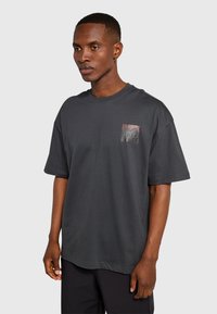 Ciemnoszary, oversize T-shirt z bawełny z małym nadrukiem kieszeni przedstawiającym panoramę miasta. Krótkie rękawy, okrągły dekolt, luźny krój.