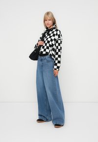 Zwart-wit geblokt cropped fleece jack, gecombineerd met lichtblauwe wijde jeans. Model houdt een zwarte schoudertas vast, staande op een witte achtergrond.