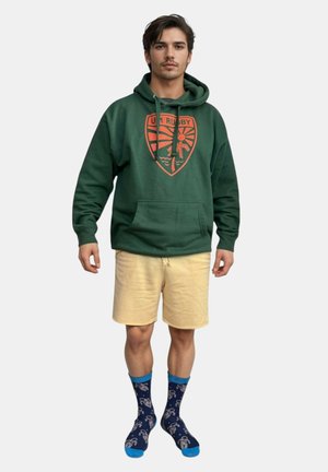 Grüne Kapuzenjacke mit orangefarbigem Grafikdesign, gelbe Shorts und blaue Socken mit Astronauten-Muster. Lässige, leichte Materialien.