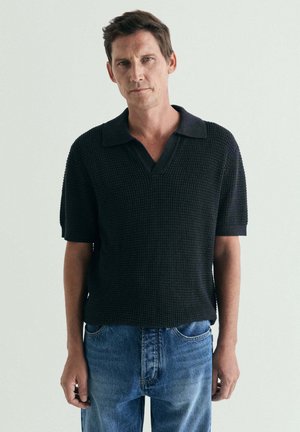 Hombre con el pelo castaño corto, vestido con una camisa tipo polo negra con textura y mangas cortas y vaqueros azules, de pie frente a un fondo claro y liso.