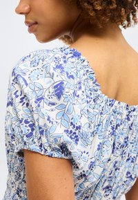 Γυναίκα που φοράει λευκό μπλουζάκι με μπλε φλοράλ σχέδιο και ραμμένο ελαστικό ντεκολτέ και μανίκια, εμφανιζόμενη από τον ώμο και τον λαιμό.