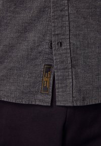 Gros plan d'une chemise grise texturée avec un bouton noir et une petite étiquette noire portant le texte jaune "ESTD 1982 STANDARD ISSUE".