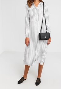 Žena v bílé midi šatech s dlouhým rukávem a vertikálním pruhem, s černou crossbody taškou a černými pletenými nazouváky na pozadí bez vzoru.