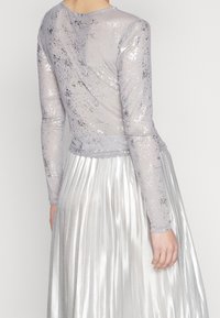 Langarmtop aus transparentem, metallisch grauem Stoff mit Glitzerelementen, kombiniert mit einem plissierten silbernen Rock, der glatte Texturen und Glanz präsentiert.