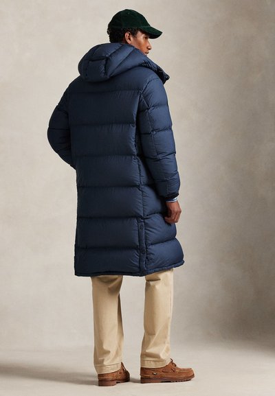 Sötétkék kapucnis téli kabát vízszintes steppeléssel és térdig érő szabással. Bézs nadrággal és barna bőr kényelmes cipővel párosítva.