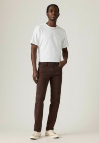 Jeans de mezclilla marrones con corte ajustado, combinados con una camiseta blanca básica y zapatillas blancas, sobre un fondo neutro.