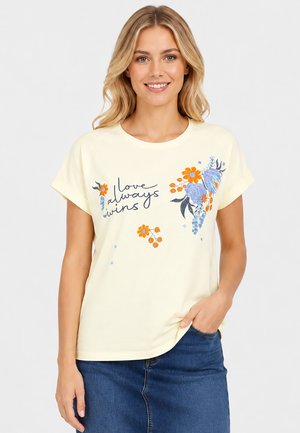 Lachende vrouw met blond haar die een lichtgele blouse draagt met tekst "love always wins" en bloemmotieven, gecombineerd met een blauwe spijkerrok.