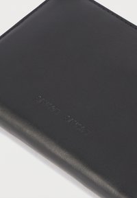 Portefeuille en cuir noir avec le nom de la marque discrètement embossé sur une surface lisse, présenté sur un fond blanc uni.