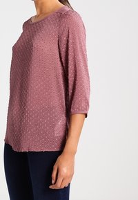 Blouse rose poussiéreux avec des manches 3/4, motifs texturés et col rond. Fabriquée en tissu léger avec une finition lisse.