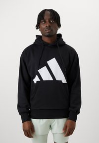 Juoda sportinė palaidinė su dideliu baltu Adidas logotipu geometriniu dizainu, su raišteliu reguliuojama kapučine ir priekinėmis kišenėmis, pagaminta iš švelnaus audinio.