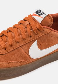 Narancssárga velúr sportcipő kerek fűzőkkel, fehér swoosh logóval és párnázott járdaszegéllyel. Jellemzője egy barna gumitalp, amelynek texturált felülete van.