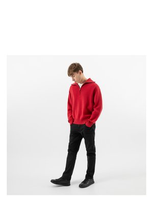 Mladý muž v červeném svetru na zip, černých kalhotách a černých teniskách, který se dívá dolů a má ruce v kapsách, na jednoduchém bílém pozadí.