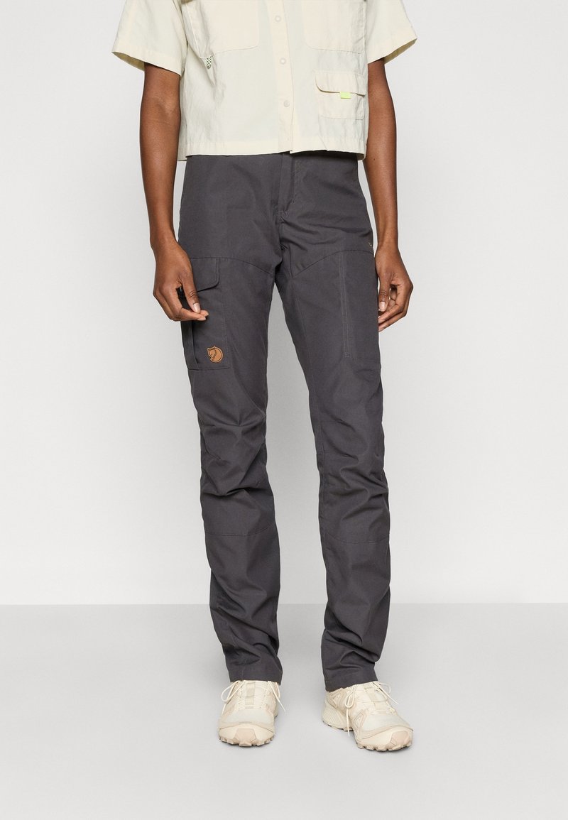 Personne portant un pantalon cargo gris foncé avec un patch logo sur la cuisse et des chaussures beiges à lacets, debout devant un fond clair uni.