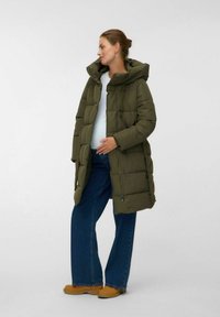 Gravid kvinde i en grøn puffjakke, hvid top, blå brede jeans og brune støvler, der står foran en ensfarvet baggrund.