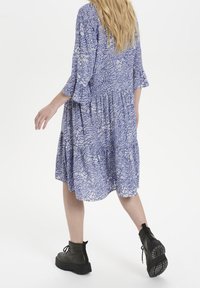 Abito a fantasia blu e bianca con maniche svasate e gonna a strati; indossato con stivaletti neri, che mette in risalto un design morbido e fluido.