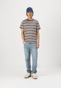 T-shirt rayé en beige, marine et marron, associé à un jean bleu clair. La tenue comprend une casquette marine et des baskets blanches avec des accents marron.