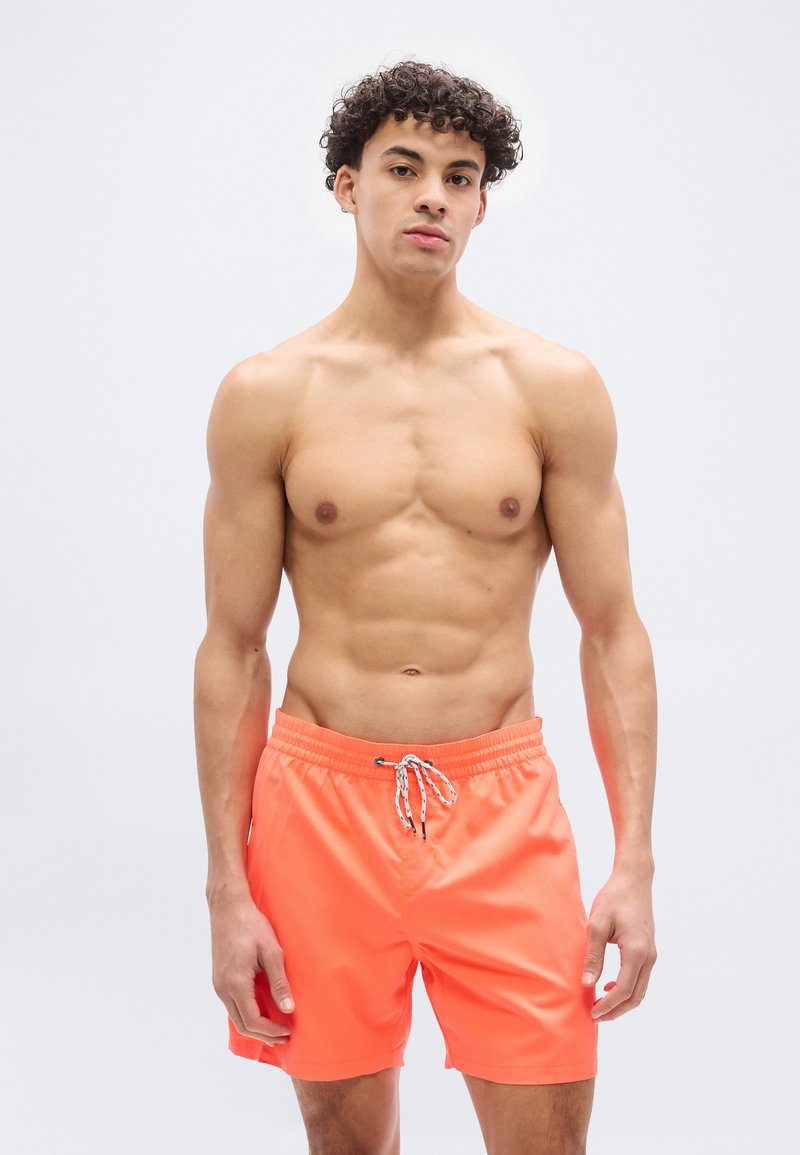 Νέος άνδρας με σγουρά μαλλιά, χωρίς πουκάμισο, φοράει φωτεινά πορτοκαλί μαγιό, στέκεται μπροστά σε απλό ανοιχτόχρωμο φόντο.