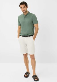 Polo verde a maniche corte con colletto a tre bottoni, pantaloni corti crema, cintura intrecciata nera e sandali neri scivolati su uno sfondo semplice.