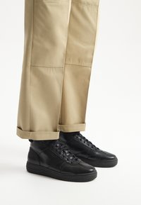 Czarne skórzane sneakersy za kostkę z teksturowanymi akcentami po bokach, zestawione z jasnobrązowymi spodniami  trzy czwarte z podwiniętymi mankietami.