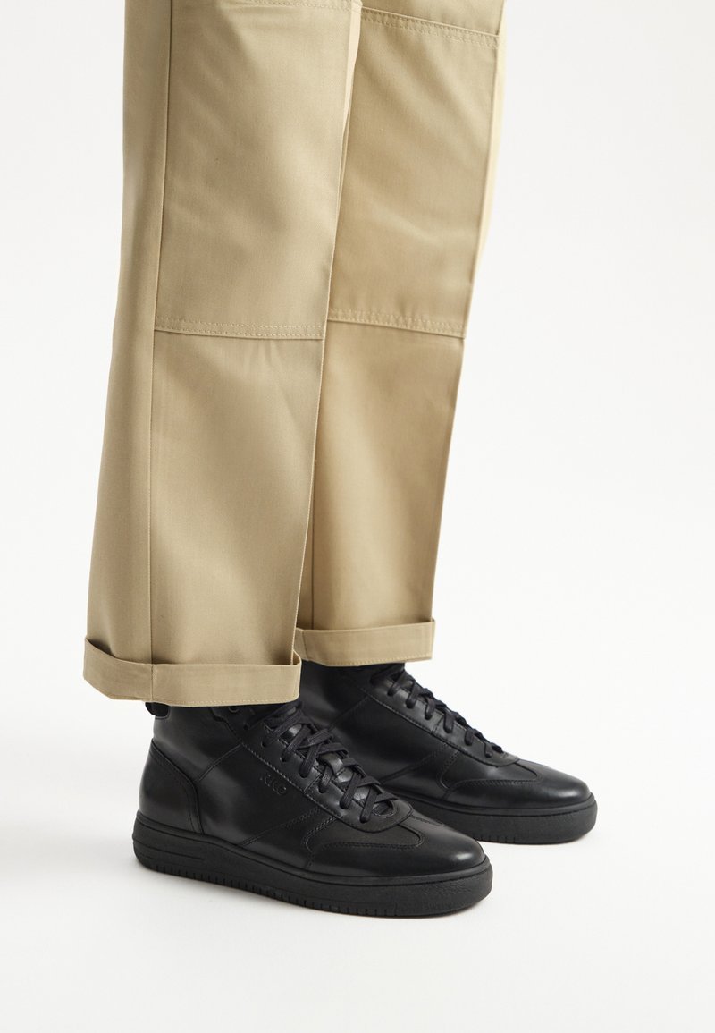 Czarne skórzane sneakersy za kostkę z teksturowanymi akcentami po bokach, zestawione z jasnobrązowymi spodniami  trzy czwarte z podwiniętymi mankietami.