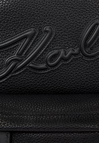 Črna teksturirana usnjena torba z izrazitim vtisnjenim napisom. Površina ima kamenčkast finiš z gladkimi poudarki v usklajeni barvi.