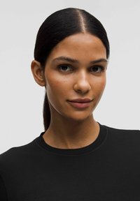 Črn, lahek top s krožnim izrezom z gladko teksturo. Model ima ravne temne lase in minimalne dodatke ter prikazuje nevtralen izraz.
