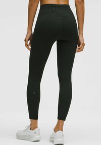 Fekete, magas derekú, rugalmas anyagból készült leggings, varrással díszítve és kis logóval, fehér sportcipővel párosítva.