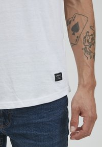 Hvid bomulds T-shirt med et sort mærke med teksten "11 PRECT" og blå denim jeans. Hånd synlig med en tatovering og et spillekortdesign.