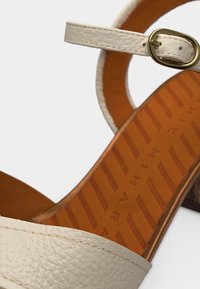 Λευκό δερμάτινο σανδάλι με λουρί στον αστράγαλο και χρυσή πόρπη, με καφέ πάτο με ανάγλυφο κείμενο και διαγώνιες γραμμές.