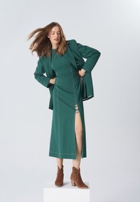 Turkusowa sukienka z bocznym rozporkiem i dekoracyjnym pierścieniem, w połączeniu z pasującą marynarką i brązowymi botkami za kostkę. Gładka faktura i wyraźnie zaznaczone szwy.