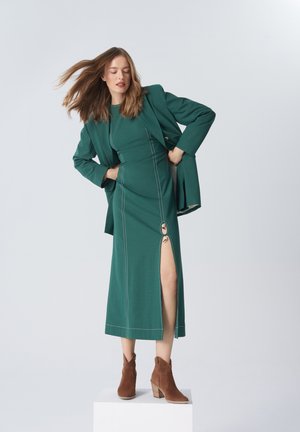 Turkusowa sukienka z bocznym rozporkiem i dekoracyjnym pierścieniem, w połączeniu z pasującą marynarką i brązowymi botkami za kostkę. Gładka faktura i wyraźnie zaznaczone szwy.