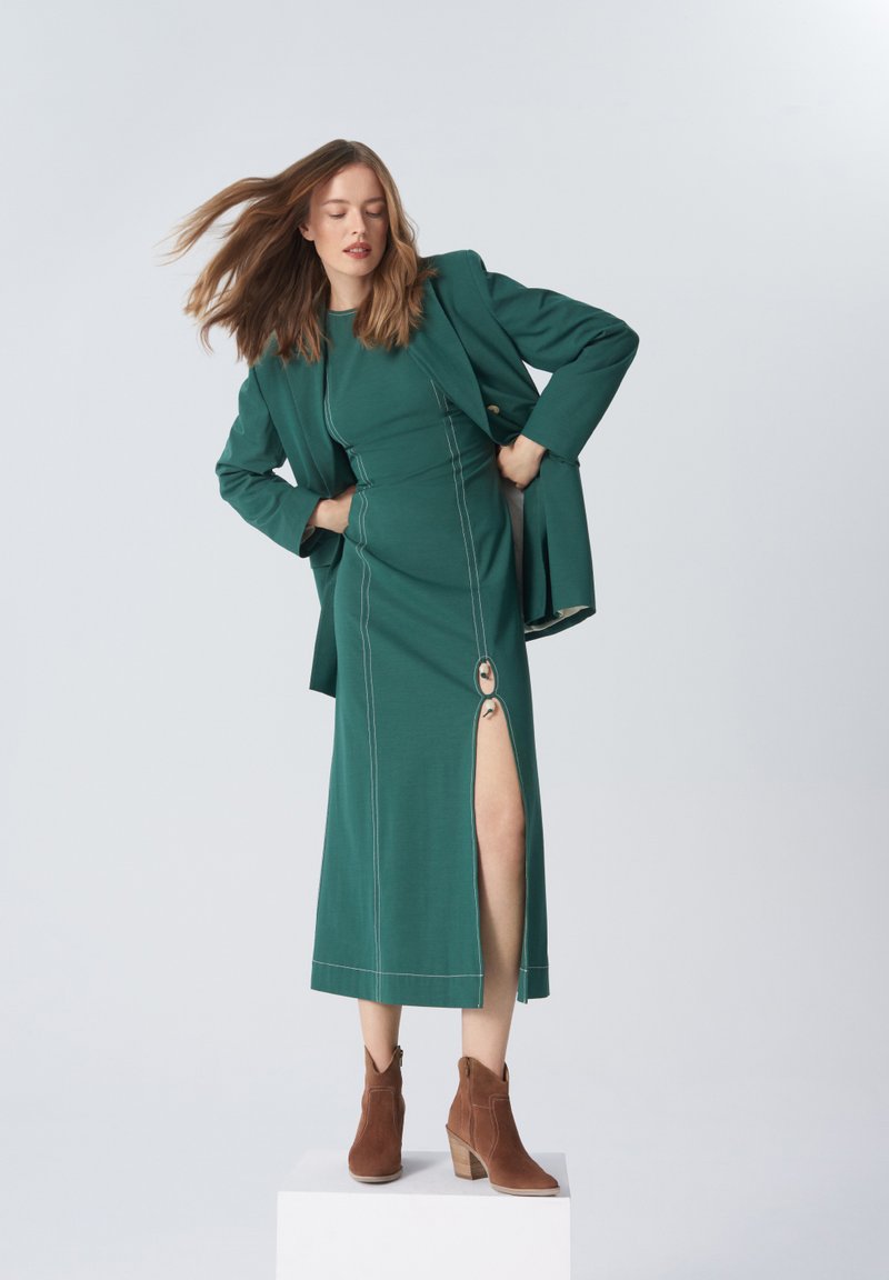 Turkusowa sukienka z bocznym rozporkiem i dekoracyjnym pierścieniem, w połączeniu z pasującą marynarką i brązowymi botkami za kostkę. Gładka faktura i wyraźnie zaznaczone szwy.