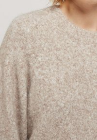 Pull en tricot beige clair avec une finition douce et texturée, dotée d'un col rond et d'une coupe ample, montré de l'épaule vers le haut.