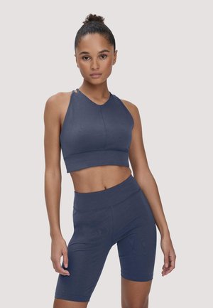 Vrouw met haar in een knot, gekleed in een mouwloos donkerblauw cropped topje en bijpassende high-waisted biker shorts, staand tegen een neutrale achtergrond.