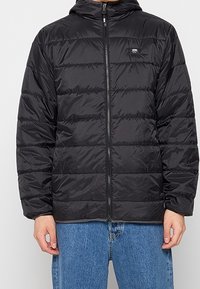 Czarna kurtka puchowa z kapturem, z zamkiem z przodu, pikowaną fakturą i logo po lewej stronie klatki piersiowej. Noszona z niebieskimi dżinsami.